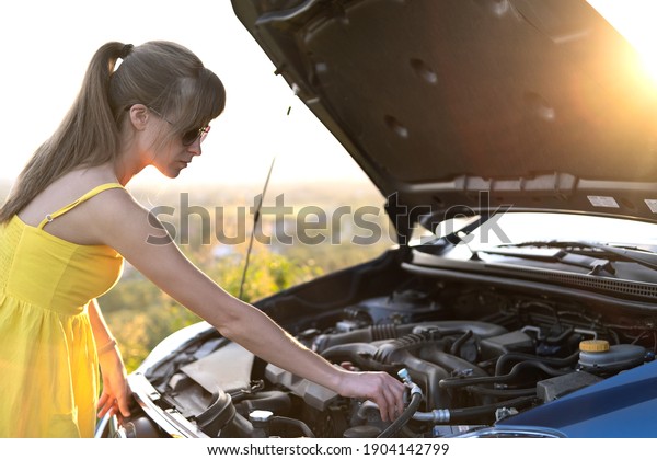 Young puzzled woman driver\
standing near her car with popped up hood looking at broken\
engine.
