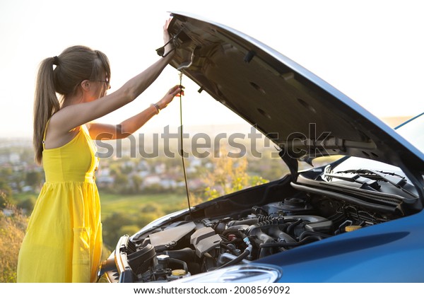 Young puzzled woman driver\
standing near her car with popped up hood looking at broken\
engine.
