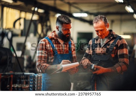 A young quality specialist checking the measures of a bearing done by a mature machinist in a factory setting. Portrait of two workers collaborating together while standing in metal processing plant.