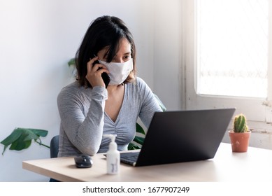 young quarantined woman talking on the phone wearing medical face mask and with hand sanitizer working from home office due to corona virus outbreak - Powered by Shutterstock