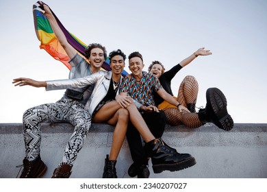 Young queer people celebrating gay pride. Group of four friends holding the rainbow pride flag while sitting together outdoors. Cheerful members of the LGBTQ+ community celebrating together. - Powered by Shutterstock