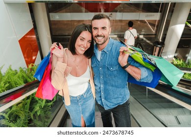 Young real couple going upstairs on the escalator of the mall carrying shopping bags and looking at camera with a smile. Customer people enjoying, smiling and walking at the steps of the store center - Powered by Shutterstock