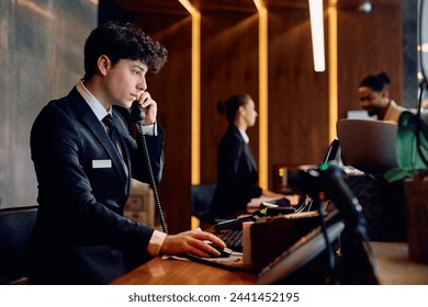 Young receptionist making a phone call while working on desktop PC at hotel reception desk.  - Powered by Shutterstock