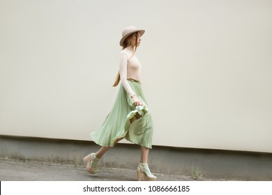 Young red hair girl in hat, dressed in beige blouse and turqoise pleats skirt, on light street backgraund.  Fashion and stylish concept.