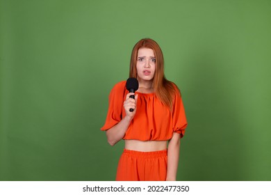 Young red hair woman in casual orange blouse on green background holding microphone worried shy to talk, confused