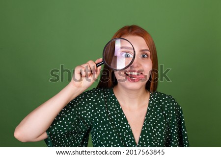 Young red hair woman on green background with magnifier playful shows  tongue