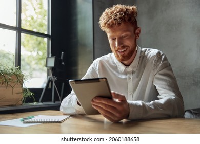 Young red haired man at wooden desk. Man in white shirt using tablet. Concept of modern successful male. Bearded stylish guy wearing formal clothes. Interior of dark room at daytime. - Powered by Shutterstock