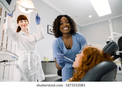 Young Red Haired School Girl During The Dental Procedure At Modern Dental Office. Female African Dentist And Her Caucasian Assistant Examinating Baby Teeth. Focus On Smiling Black Woman Dentist