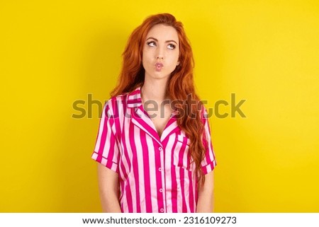 Young red haired woman wearing pink pyjama over yellow studio background making fish face with lips, crazy and comical gesture. Funny expression.