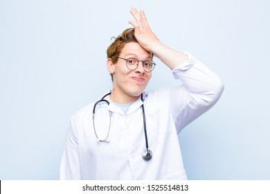 Young Red Head Doctor Raising Palm To Forehead Thinking Oops, After Making A Stupid Mistake Or Remembering, Feeling Dumb Against Blue Wall