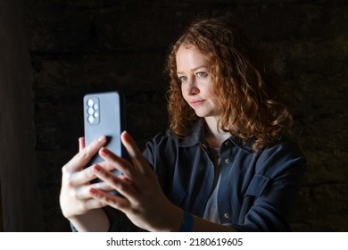 Young Red Head Female Smiles Taking Selfies For Social Media. Influencer Takes Portrait With Smartphone. Natural Light From Window Against Dark Background. Girl Unlocks Phone With Face Recognition.