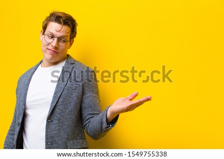 young red head man feeling happy and smiling casually, looking to an object or concept held on the hand on the side against orange background
