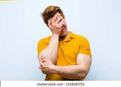 young red head man looking stressed, ashamed or upset, with a headache, covering face with hand against blue wall