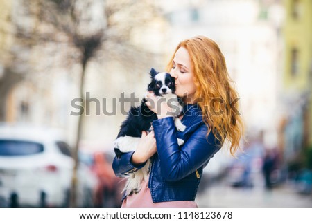 A young redhaired Caucasian woman with freckles holds and kisses, embracing black and white shaggy dog of Chihuahua breed. girl dressed in blue leather jacket, stands on street in spring in Europe.