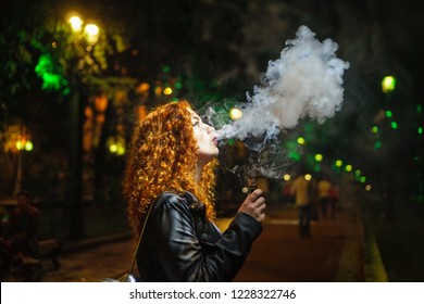 Young red-haired girl dreamily smokes an electronic cigarette. She walks alone on streets of night city. Non-nicotine cigarette. Wet asphalt after rain. - Powered by Shutterstock