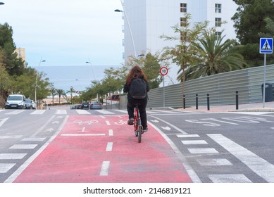 Young Red-haired Girl, Real People, Riding Red Bike With Black Backpack Rides Along A Red Bike Path Towards The Beach In Benidorm, Spain. Very Long Shot.