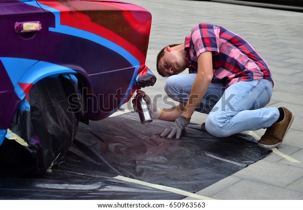 A young red-haired graffiti artist paints a new
colorful graffiti on the car. Photo of the process of drawing a
graffiti on a car close-up. The concept of street art and illegal
vandalism