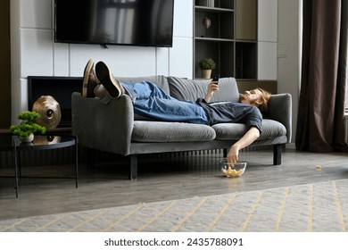 A young red-haired guy is lying on the sofa and eating corn flakes, scrolling through social networks on his phone