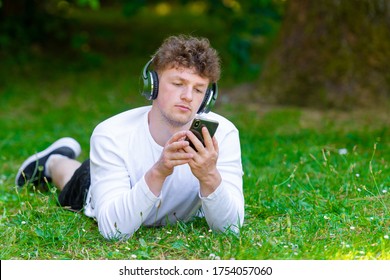 Young Red-haired Man In Headphones Lies On The Green Grass Looking At The Phone