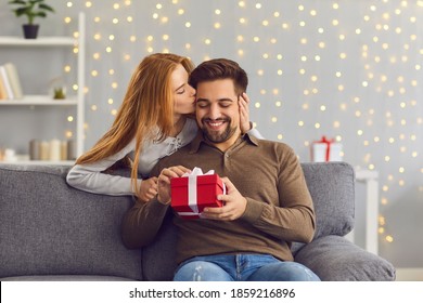 Young red-haired woman from behind hugs and kisses her beloved husband giving him a gift on this special day. Concept of gifts for birthdays, Christmas, Valentine's Day and Men's Day.