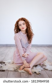 A young red-haired woman with blue eyes, dressed in a dress with polka dots, sits on a rug made of animal skin against the background of a white room.