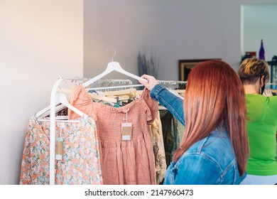 Young Red-haired Woman Buying Clothes From A Rack, In A Small Fashion Shop. Shopping Concept.