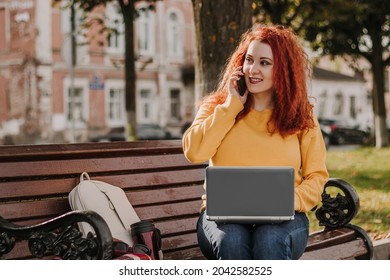 Young red-haired woman freelancer works in park using laptop. Modern businesswoman talking on phone. Online work on the Internet.