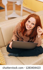 Young red-haired woman in knitted sweater sitting with her legs crossed on couch with thin laptop computer in her hands, looking at camera and smiling. Portrait from high angle