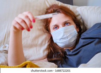 Young Red-haired Woman Looks At A Thermometer While Lying In Bed. Sick Girl Lies At Home In A Medical Mask