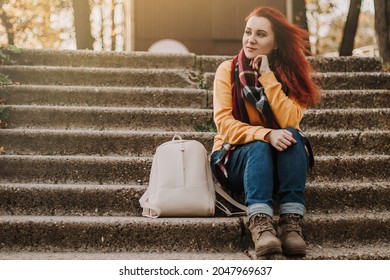 Young Red-haired Woman Sits On Stairs In Park. Lady In Yellow Sweater Is Enjoying Sunny Day. Urban Tourism. Lifestyle, Copy Space.