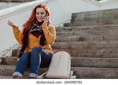 Young Red-haired Woman Sitting On Stairsand Talking On Phone. Lady In Yellow Sweater Uses Smartphone On Sunny Day. Lifestyle, Copy Space.