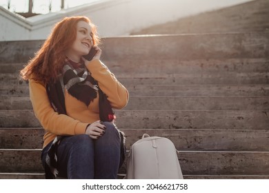 Young Red-haired Woman Sitting On Stairsand Talking On Phone. Lady In Yellow Sweater Uses Smartphone On Sunny Day. Lifestyle, Copy Space.