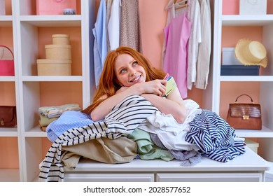 young redhead attractive caucasian woman happy positive smile hold credit card buy purchase clothes shopper sit behind table with outfits at home in cozy bright room, shopaholic. consumerism concept