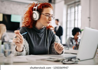 Young Redhead Businesswoman Enjoying In Music Over Headphones While Reading An E-mail On Computer At Work. Here Are People In The Background. 