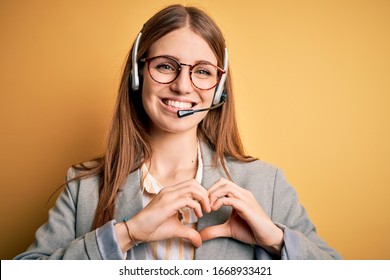 Young redhead call center agent woman overworked wearing glasses using headset smiling in love showing heart symbol and shape with hands. Romantic concept.