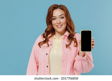 Young Redhead Chubby Overweight Woman 30s With Curly Hair Wear Pink Shirt Casual Clothes Using Mobile Cell Phone With Blank Screen Area Mock Up Show Thumb Up Gesture Isolated On Pastel Blue Background