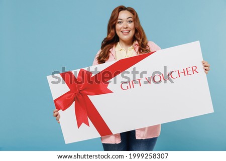 Young redhead chubby overweight woman 30s with curly hair wear casual pink shirt hold big gift certificate coupon voucher card for store isolated on pastel blue background People lifestyle concept.