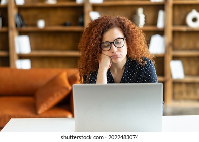 Young Redhead Curly Woman Using Laptop Sitting In The Office And Feels Bored, Fatigue, Lack Of Motivation And Energy, Female Employee Tired From Not Interesting And Monotonous Work