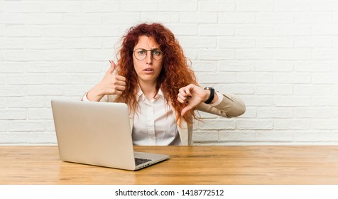 Young redhead curly woman working with her laptop showing thumbs up and thumbs down, difficult choose concept