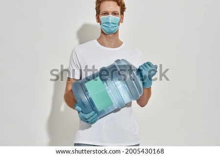 Young redhead deliveryman holds a huge bottle of water with both hands in front of him, his eyes are smiling, wearing white T-shirt ,isolated over the white wall