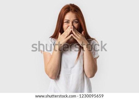 Young redhead European female isolated on white background dressed in white t-shirt, covering nose with hands experiencing stench and bad smell