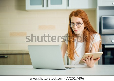young redhead female entrepreneur working online with digital tablet and laptop while sitting at table in her home office