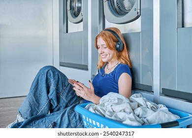 Young redhead girl in a public laundry sitting on the floor with a laundry basket next to her listening to music while waiting for her clothes to be washed - Powered by Shutterstock