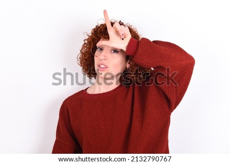 young redhead girl wearing red sweater over white background making fun of people with fingers on forehead doing loser gesture mocking and insulting.
