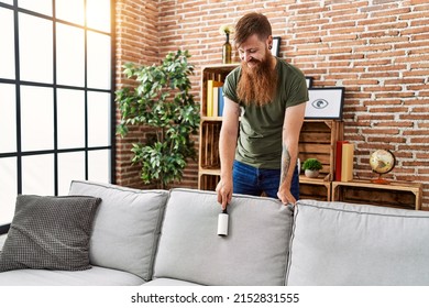 Young Redhead Man Cleaning Sofa Using Pet Hair Roller At Home