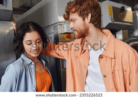 young redhead man flirting with smiling asian girlfriend near washing machine in public laundry