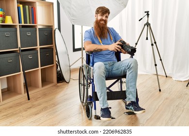 Young redhead man photographer sitting on wheelchair using professional camera at photograpy studio