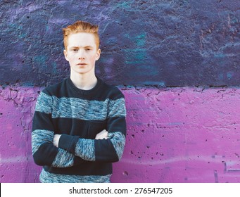 Young redhead man in a sweater and jeans standing next to purple wall with folded arms