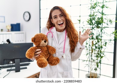 Young redhead pedriatician woman holding teddy bear celebrating achievement with happy smile and winner expression with raised hand 