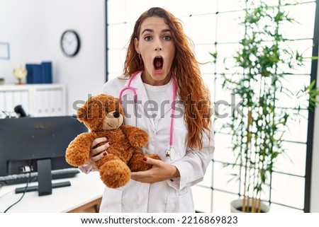 Young redhead pedriatician woman holding teddy bear scared and amazed with open mouth for surprise, disbelief face 